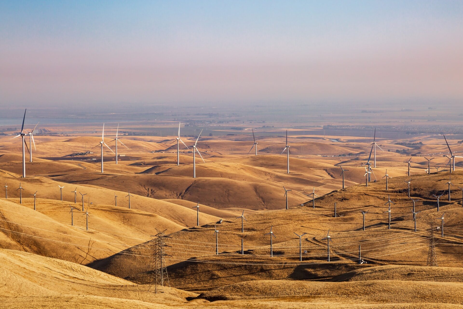 Numerous wind turbines stand on golden, rolling hills under a hazy sky, generating renewable energy across a vast, dry landscape. Power lines run through the scene, showing the transmission of electricity.