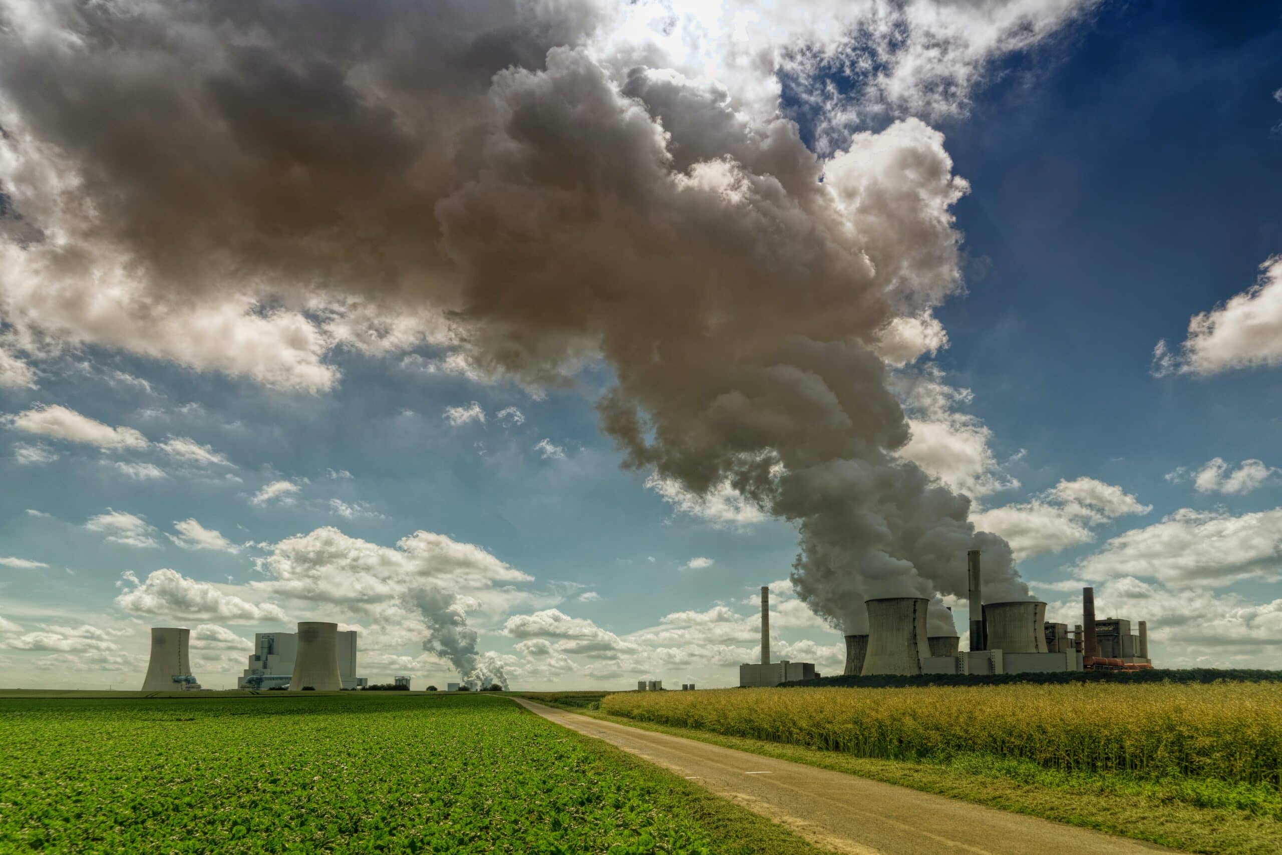 A large industrial power plant emits thick clouds of smoke into a partly cloudy sky, with green fields and a dirt road in the foreground.