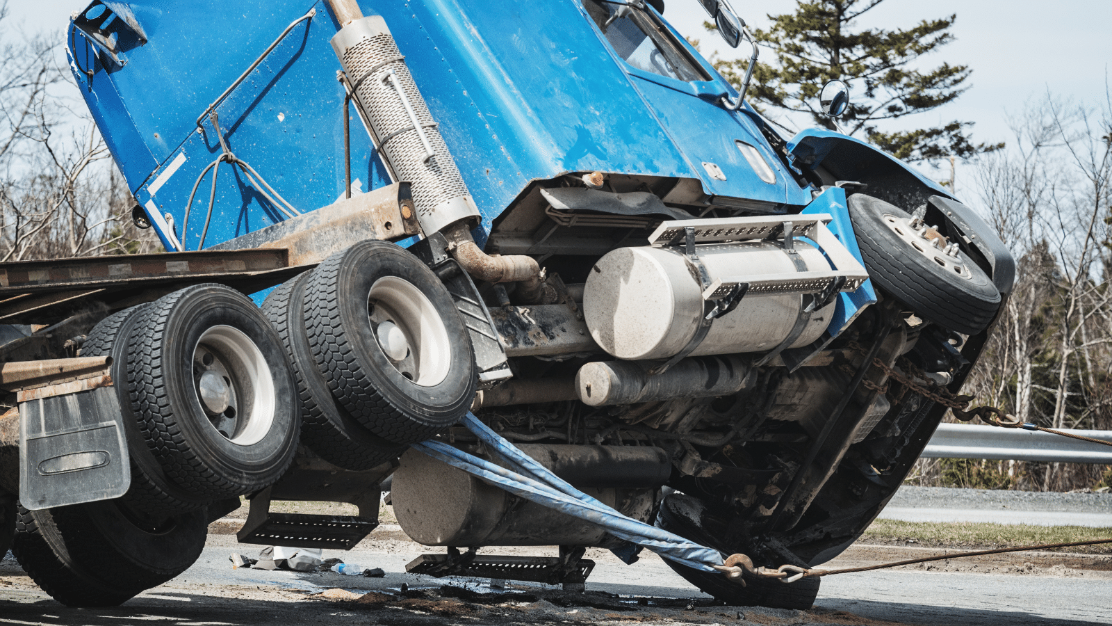 A blue semi-truck cab is tilted and partially lifted off the ground, secured by heavy straps attached to a tow truck, after an apparent accident on a road with trees in the background.