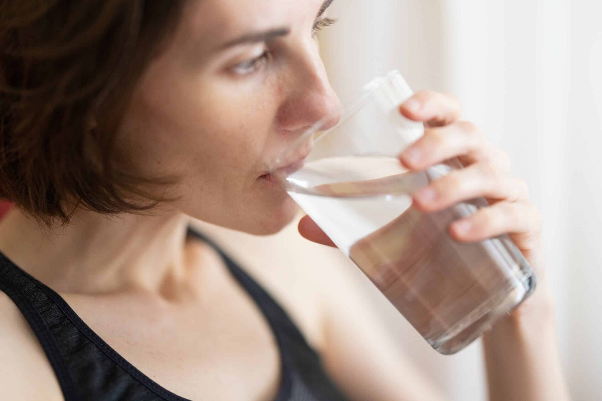 A person with short brown hair is drinking a glass of water, wearing a dark sleeveless top, and looking relaxed in a bright indoor setting.