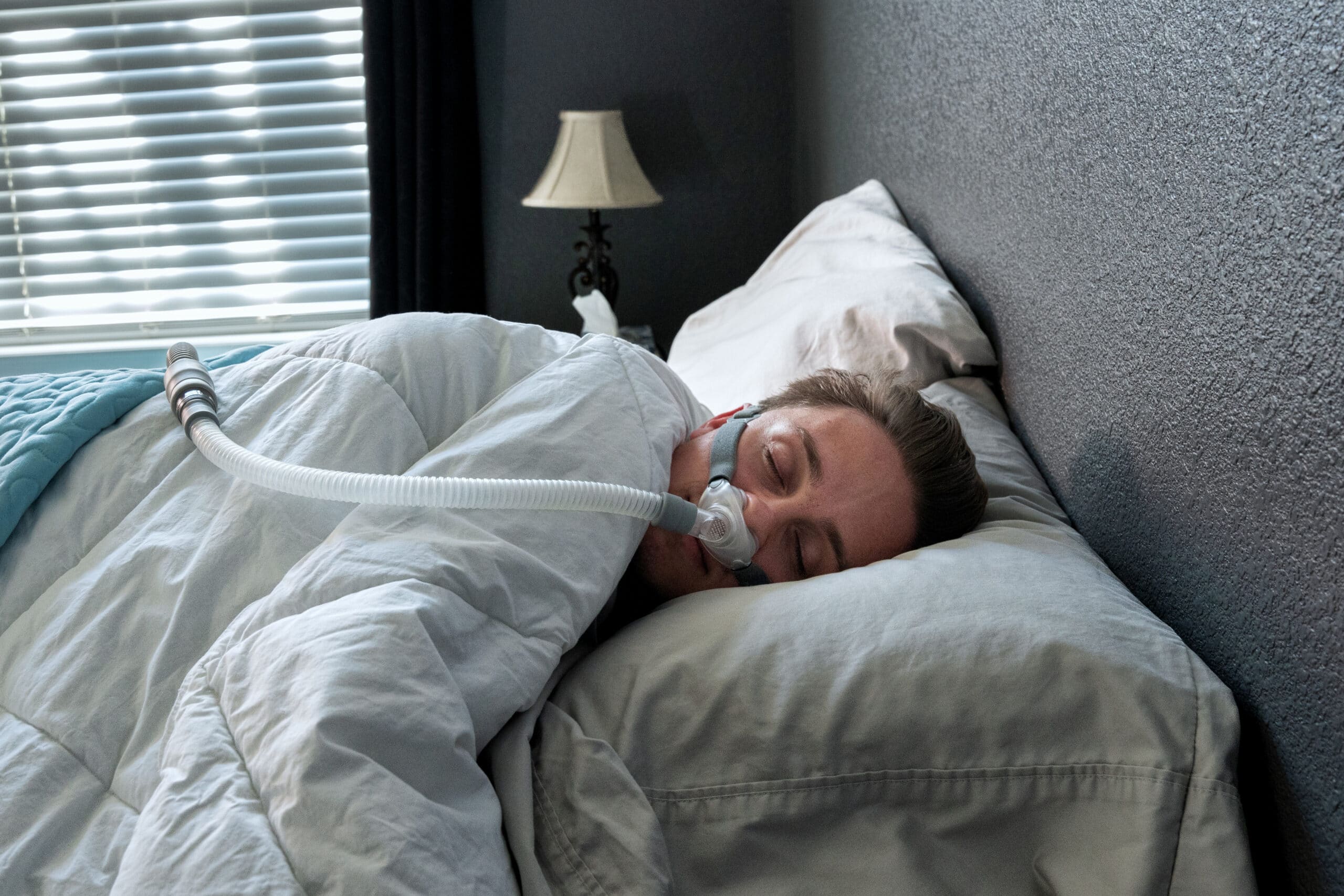 A person sleeps on their side in bed, covered with a white blanket, wearing a CPAP mask connected to a hose. Soft light comes through window blinds, and a small lamp sits on a nightstand nearby.