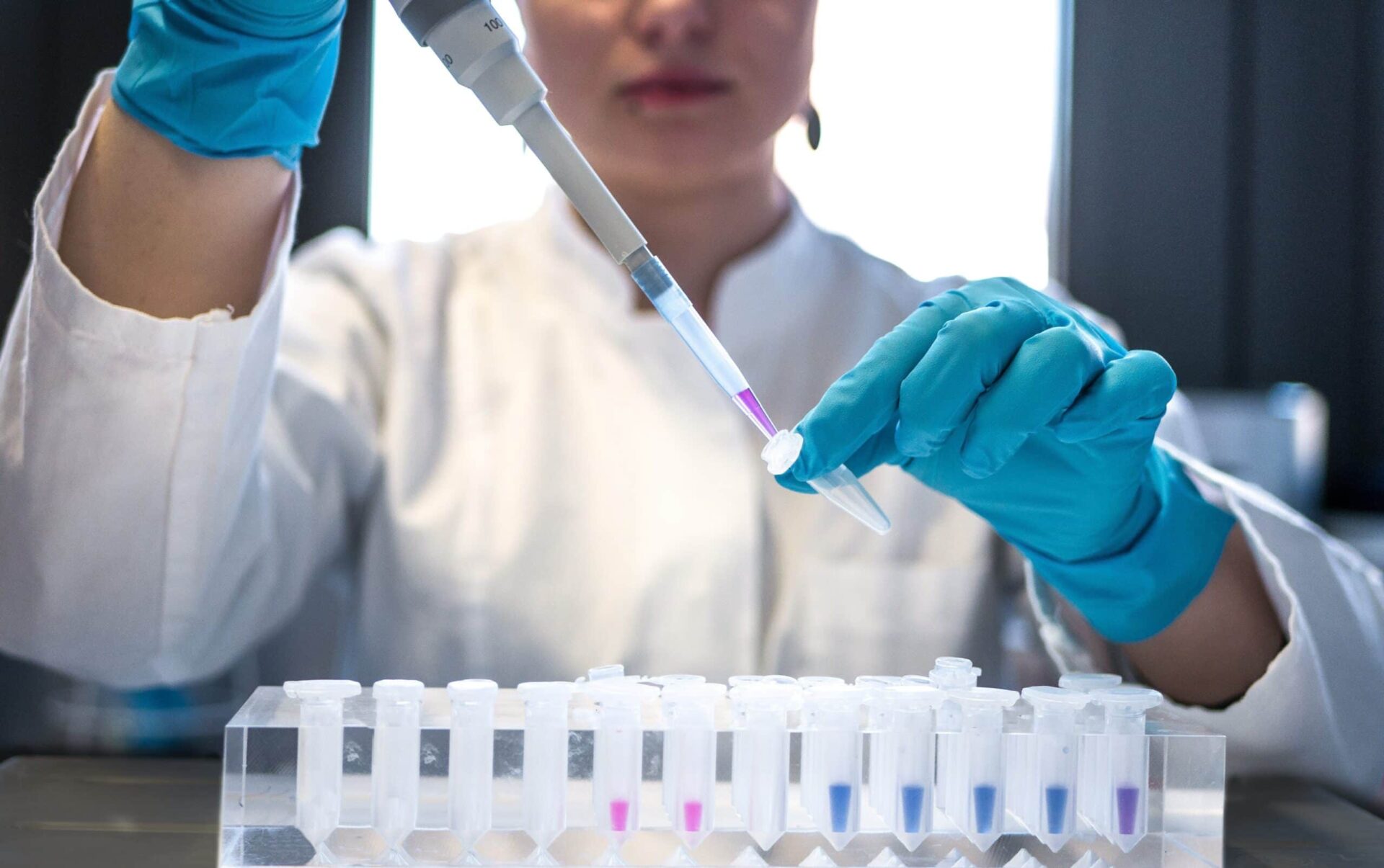 A scientist in a white lab coat and blue gloves uses a pipette to transfer liquid into a small vial, with several other vials arranged in a rack on the table.