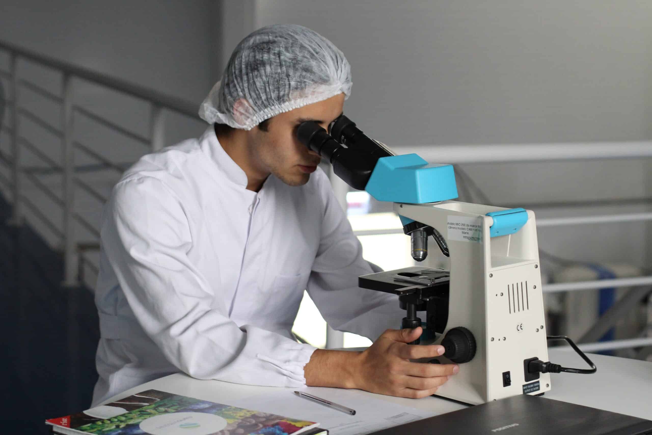 A person wearing a white lab coat and hair cover looks through a microscope at a lab table, with documents and a pen nearby.