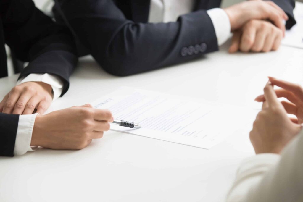 Three people in business attire sit at a white table, discussing and pointing at a document with a pen, suggesting a formal meeting or contract review. Their faces are not visible.