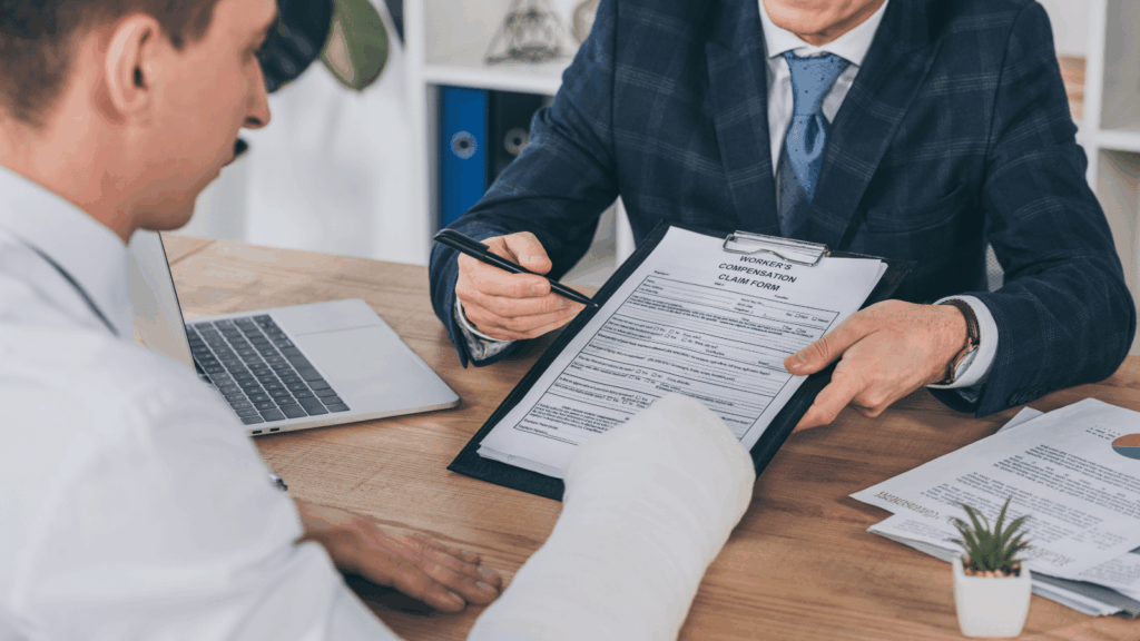 A person with a bandaged arm sits at a desk across from another person in a suit, who is pointing at a clipboard holding an insurance compensation claim form.
