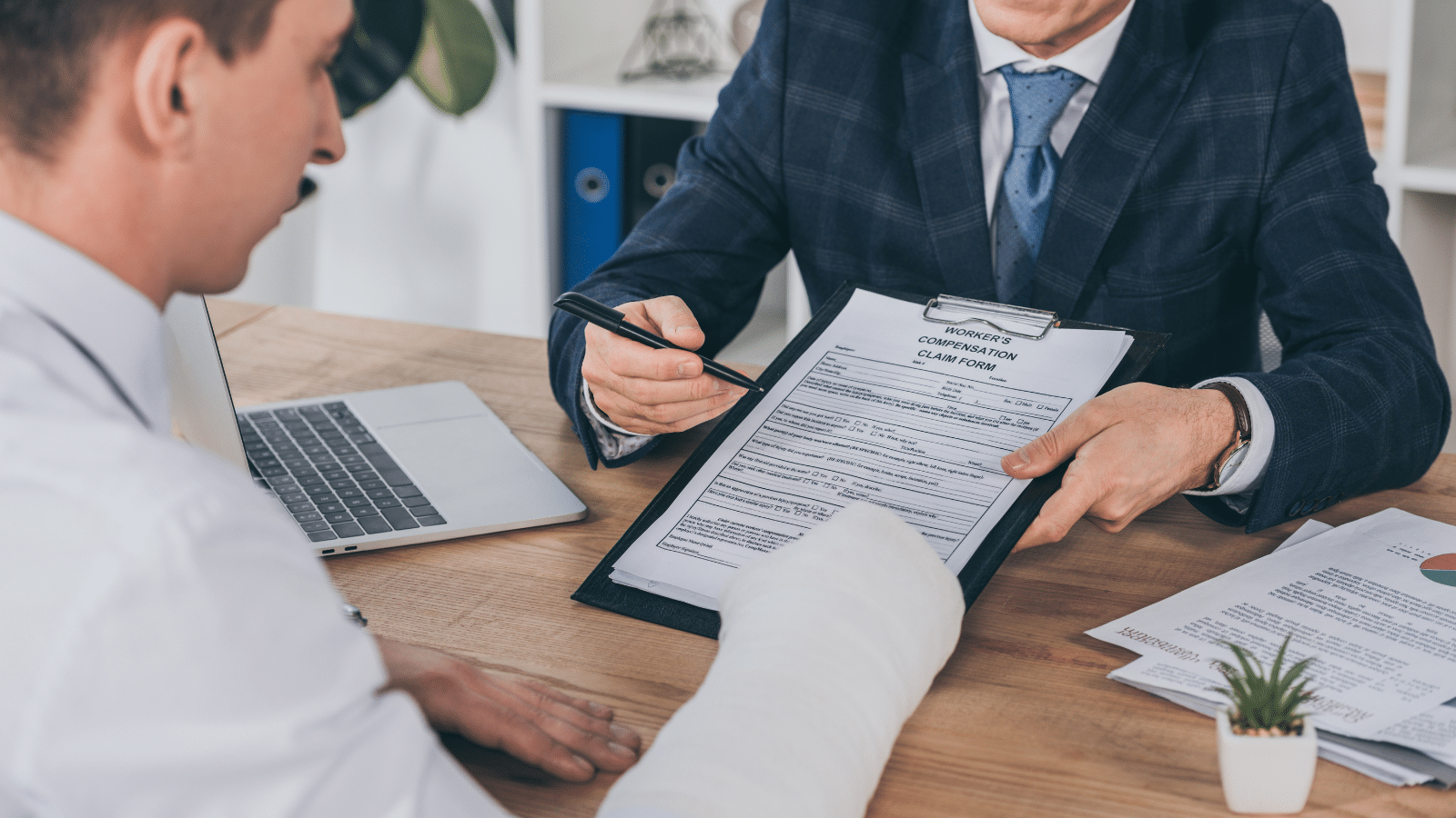 A person with a bandaged arm sits at a desk across from another person in a suit, who is pointing at a clipboard holding an insurance compensation claim form.