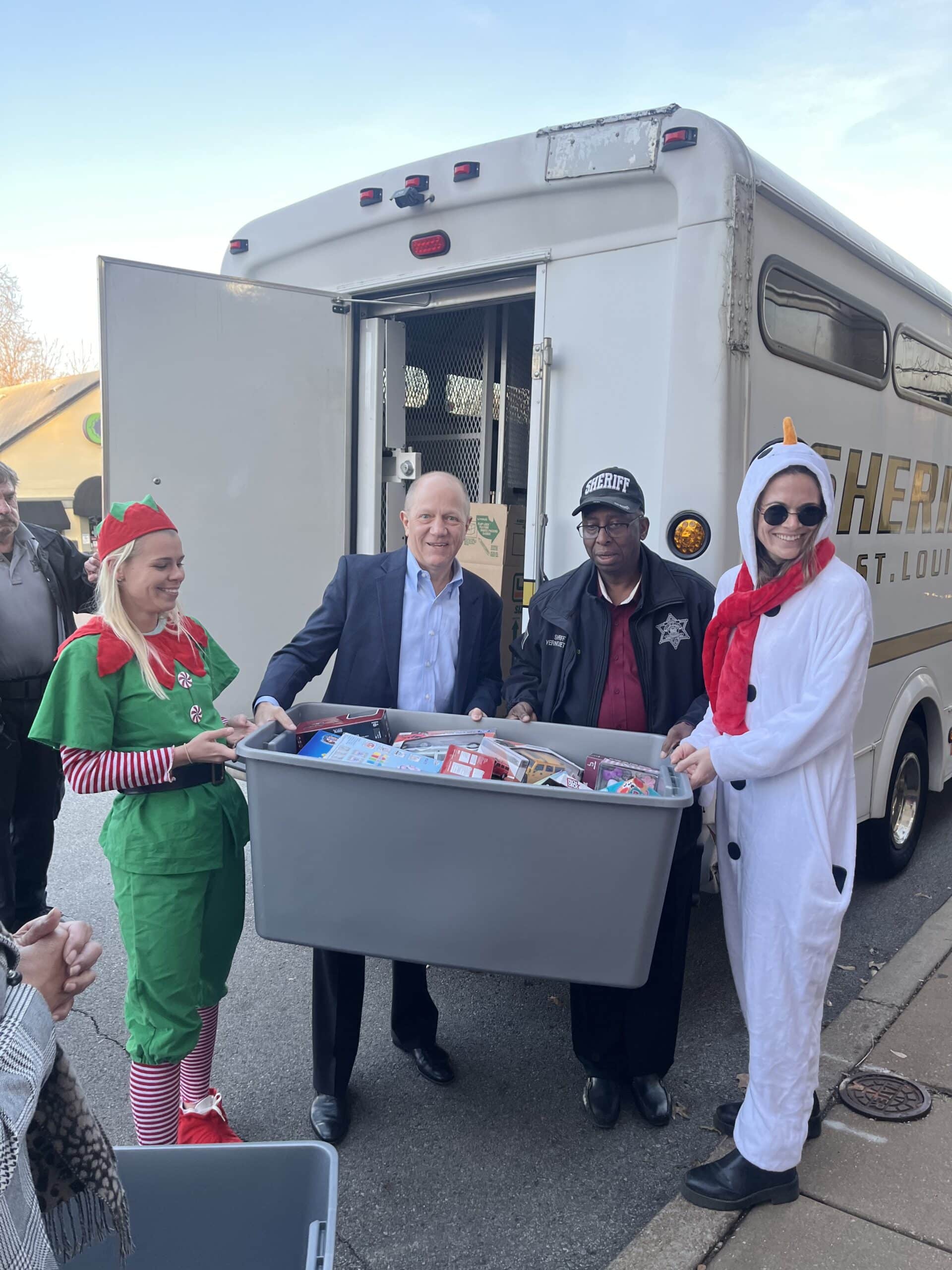 Four people, including an elf and someone in a snowman costume, stand by a sheriffs van holding a large bin filled with toys and gifts, participating in a holiday charity event.