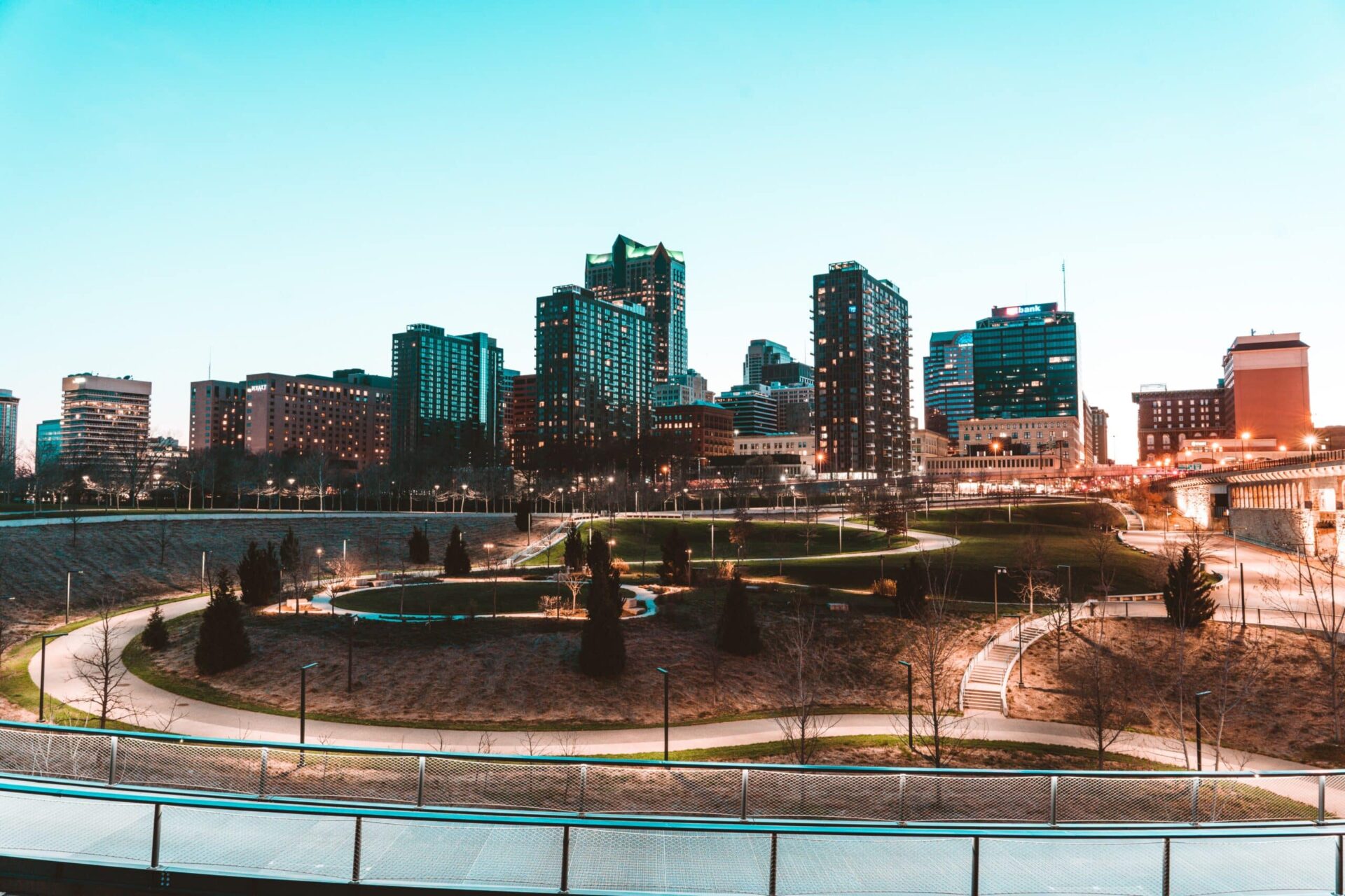 A city skyline at dusk with tall buildings in the background and a large park with winding pathways, green grass, and scattered trees in the foreground. The sky is clear and light blue.