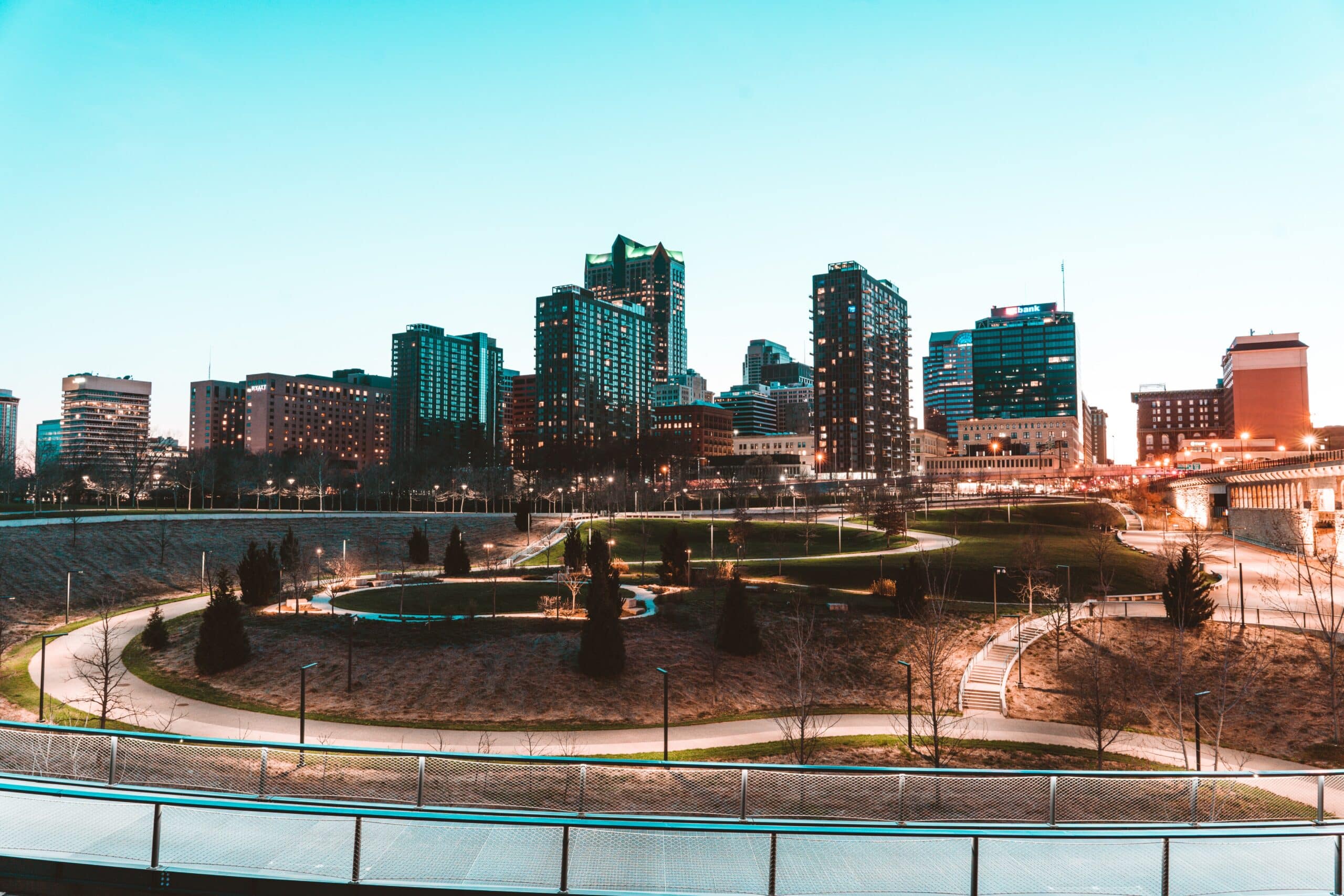 A city skyline at dusk with tall buildings in the background and a large park with winding pathways, green grass, and scattered trees in the foreground. The sky is clear and light blue.