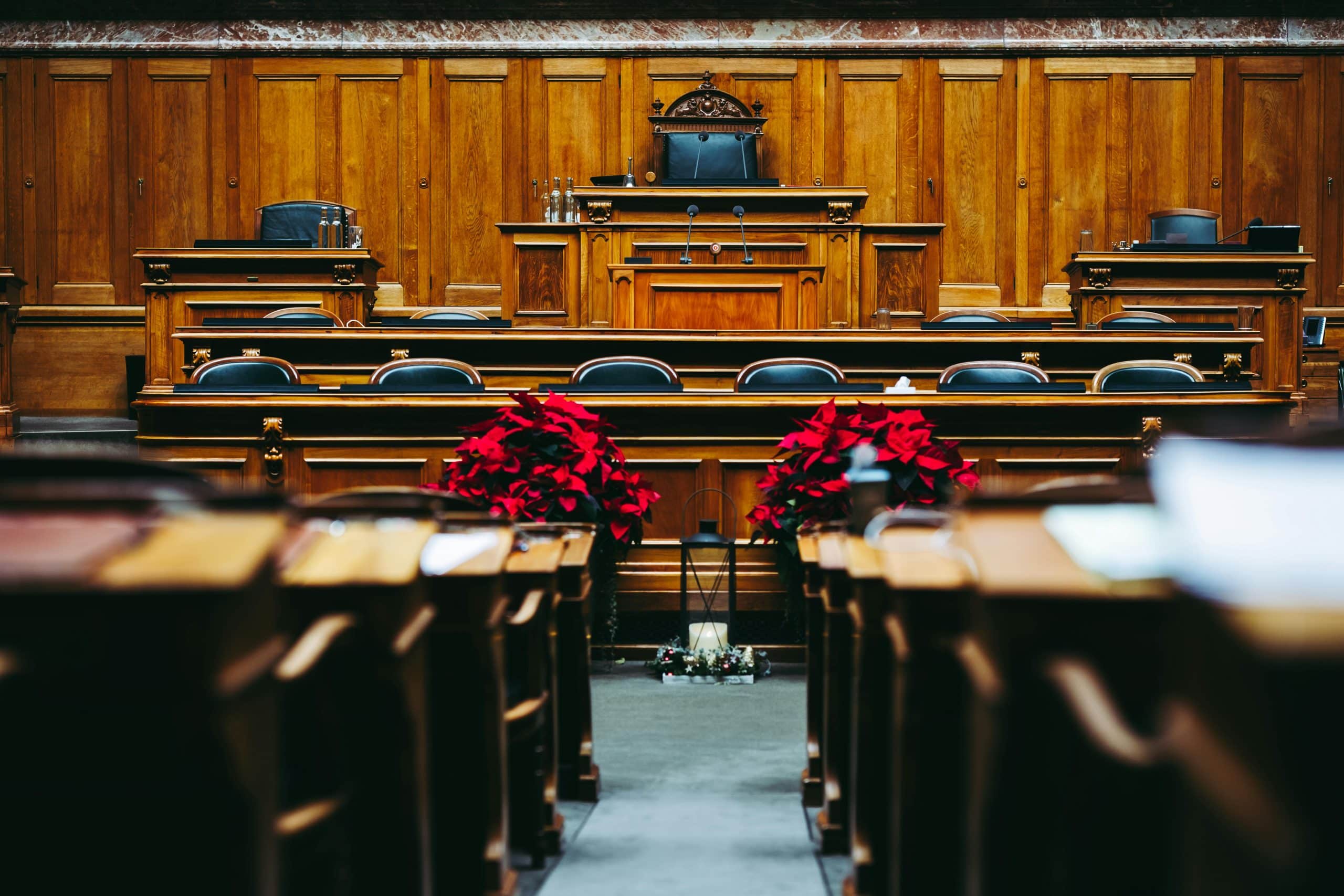 Empty wooden courtroom or legislative chamber with rows of desks, a judge’s or speaker’s chair at the front, and red flower arrangements placed in the center aisle.