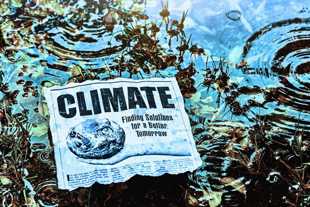 A newspaper with the headline CLIMATE: Finding Solutions for a Better Tomorrow lies partially submerged in water, surrounded by grass and ripples, suggesting themes of climate change and flooding.