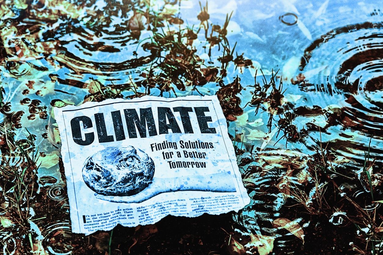 A newspaper with the headline CLIMATE: Finding Solutions for a Better Tomorrow lies partially submerged in water, surrounded by grass and ripples, suggesting themes of climate change and flooding.