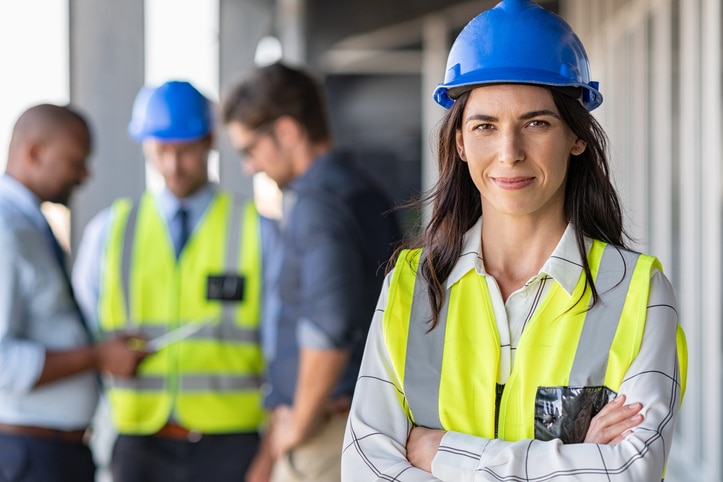 Portrait of woman engineer at building site looking at camera with copy space. Mature construction manager standing in yellow safety vest and blue hardhat with crossed arms. Successful confident architect at construction site with team discussing in background.