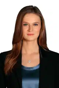Kayla Ferrel with long brown hair wearing a black blazer over a blue top, accessorized with a pendant necklace, poses against a plain white background.