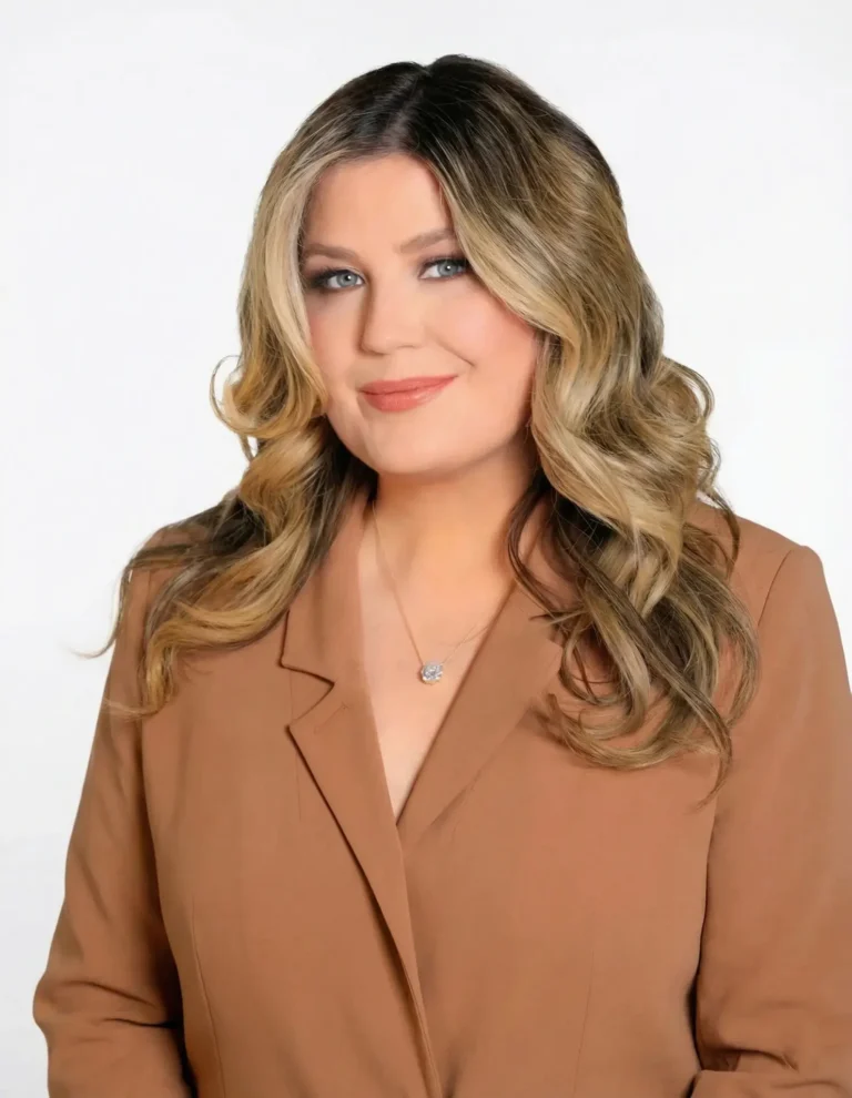 A woman with long, wavy blonde hair wears a tan blazer and a necklace, smiling slightly against a plain white background.