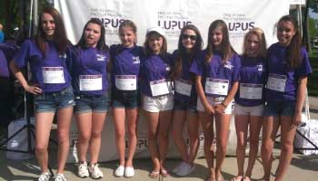 Eight young women wearing matching purple shirts and name tags stand in a line, smiling in front of a Lupus Foundation event banner outdoors on a sunny day.
