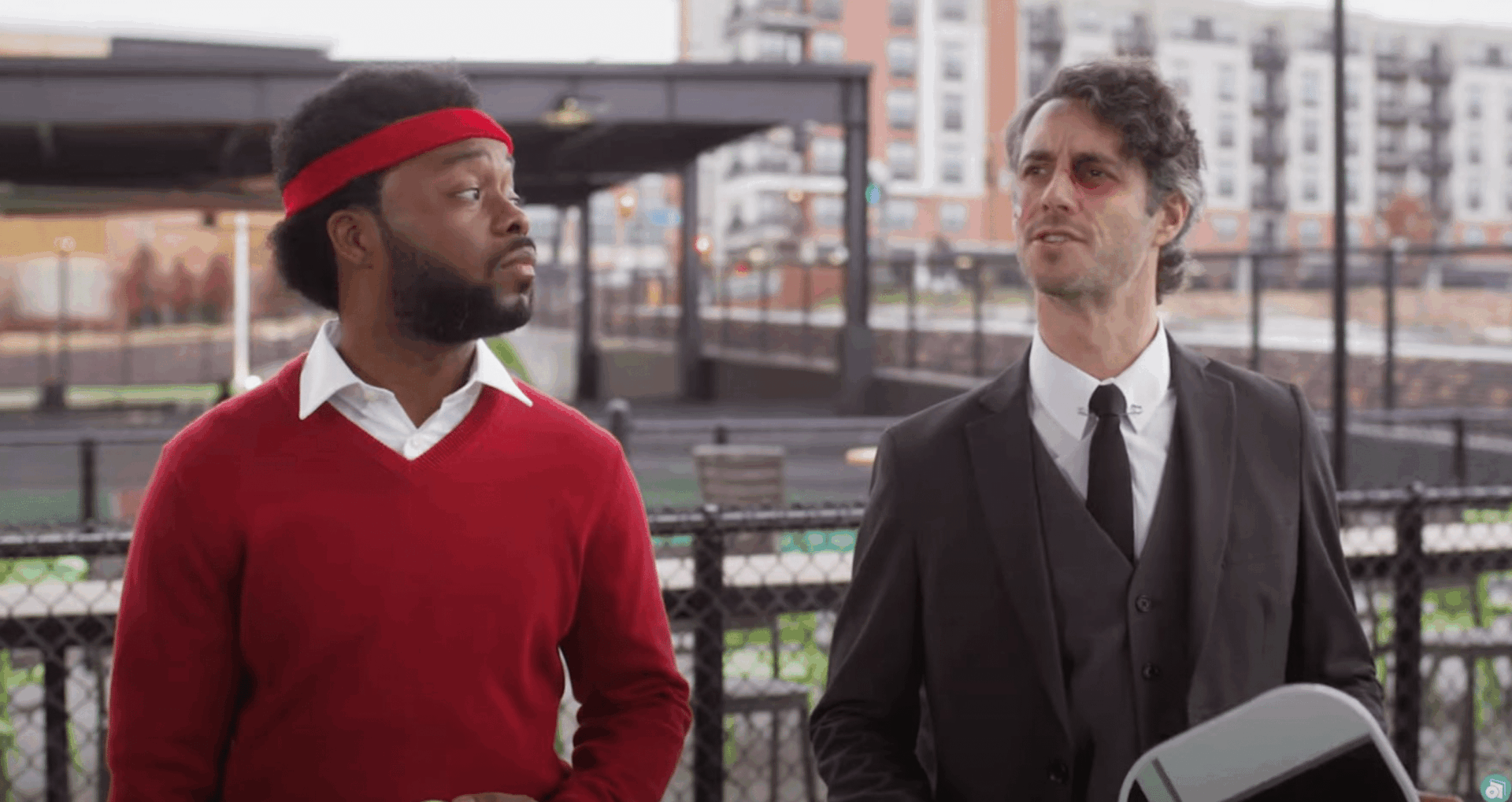 Two men stand outdoors by a fence; one wears a red sweater and headband, while the other is in a black suit and tie with a bruised face. Apartment buildings and a park are visible in the background.
