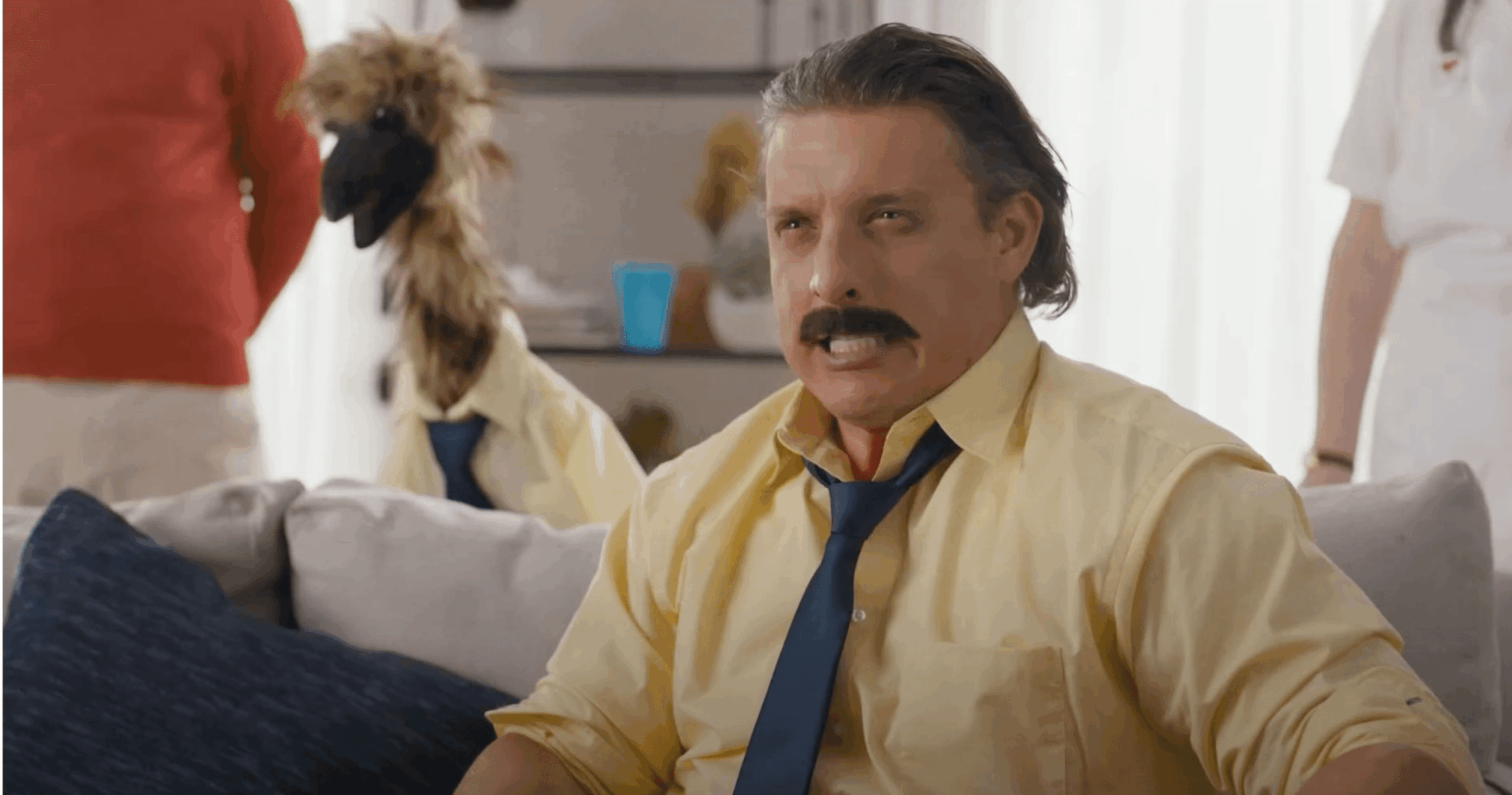 A man wearing a yellow shirt, blue tie, and fake mustache sits on a couch, making a serious face. Behind him, an emu wearing a matching yellow shirt and blue tie stands upright.