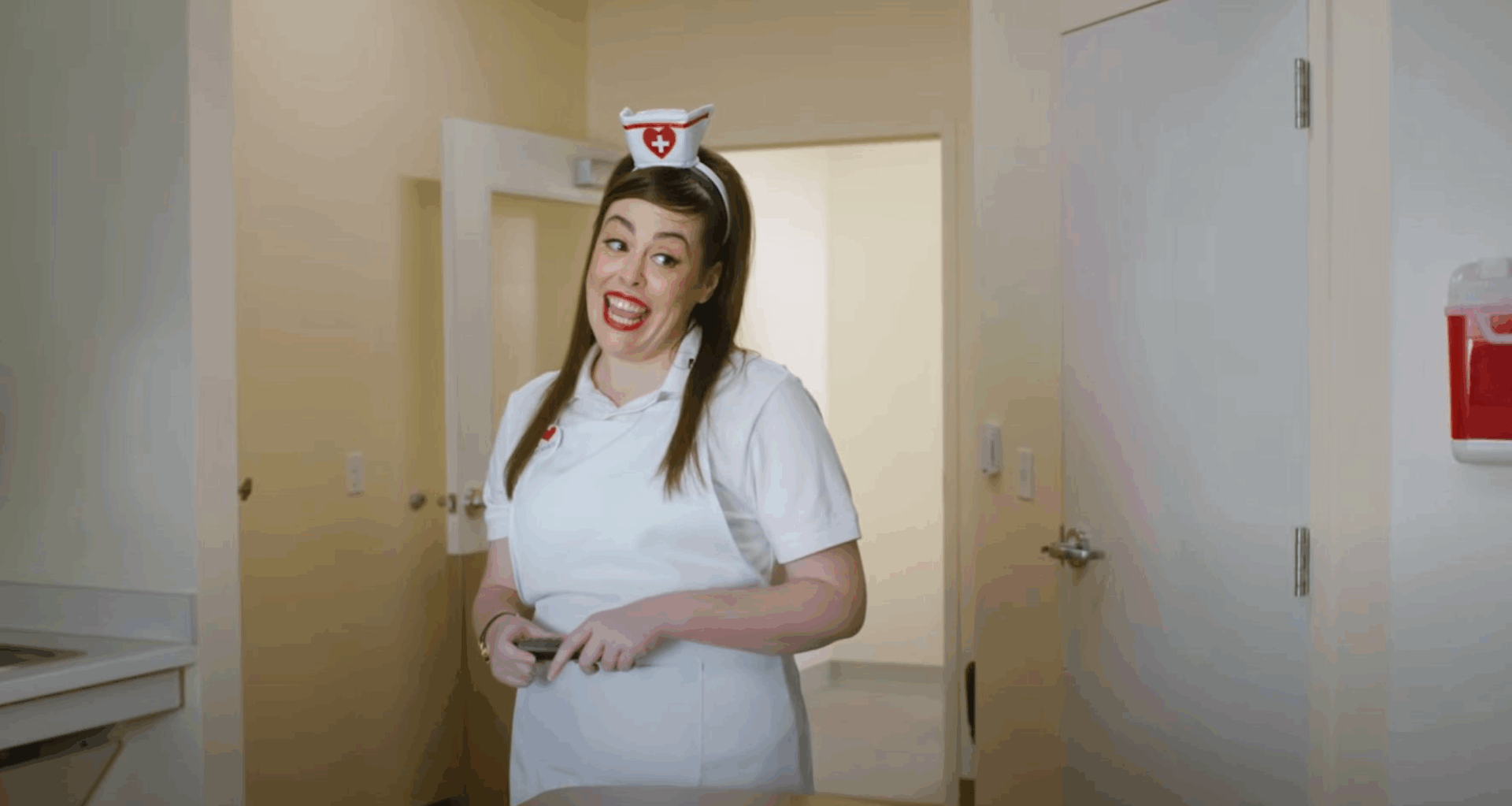 A woman dressed as a nurse in a white uniform and hat stands smiling in a brightly lit room, holding a clipboard. There is an open door and a red box on the wall behind her.
