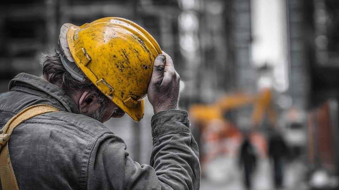 A construction worker wearing a yellow hard hat and dusty jacket stands with his head bowed, hand touching the helmet, on an industrial site with blurred figures and machinery in the background.
