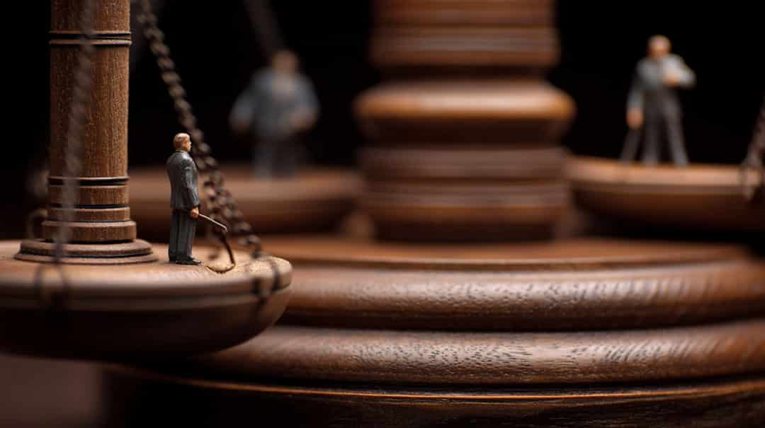 Close-up of a wooden balance scale with small figurines of people standing on its pans, symbolizing justice and fairness, with the scales and figurines in sharp focus and a blurred background.