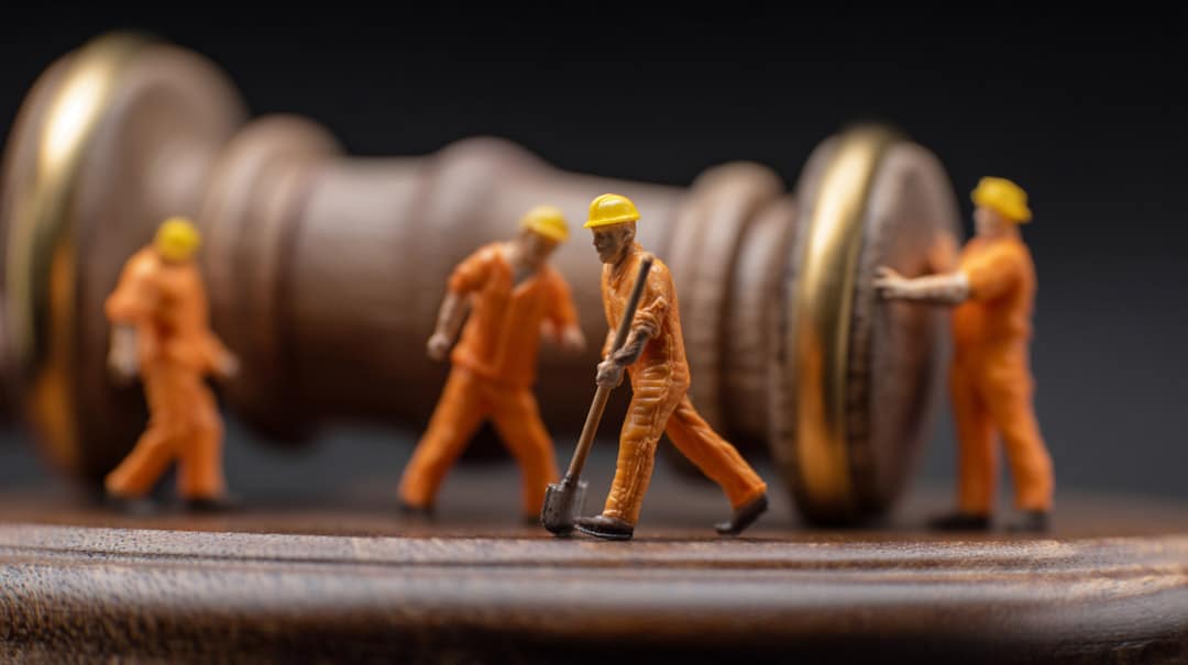 Miniature construction workers in orange uniforms and yellow helmets stand and work on a wooden gavel, symbolizing labor and justice, against a dark background.