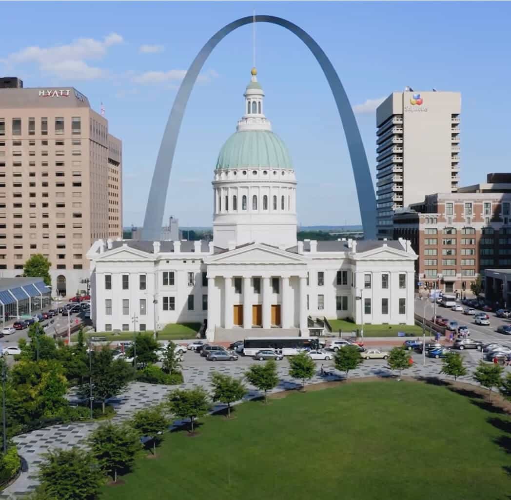 A white domed historic courthouse stands in front of the Gateway Arch in St. Louis, Missouri, surrounded by city buildings and green lawns under a clear blue sky.