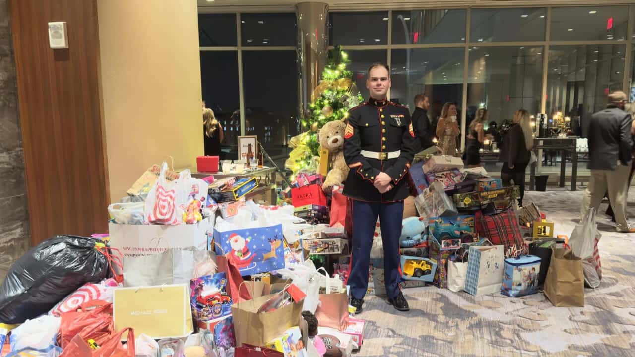 A person in a military dress uniform stands in front of a decorated Christmas tree, surrounded by piles of donated toys and gift bags in a hotel lobby. Other people and festive decorations are visible in the background.