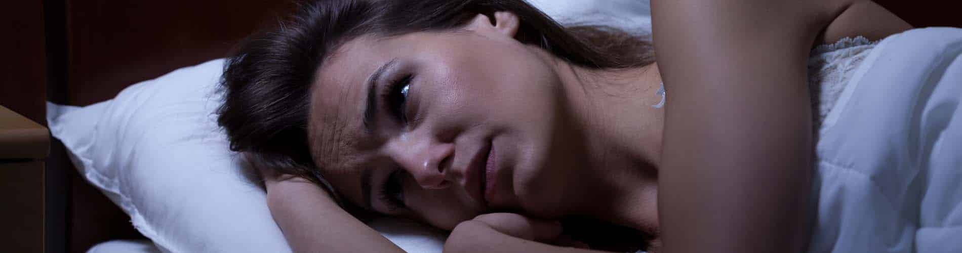 A woman lies in bed at night, resting her head on a pillow, looking worried or anxious as she stares off into the distance. The room is dimly lit, creating a somber mood.