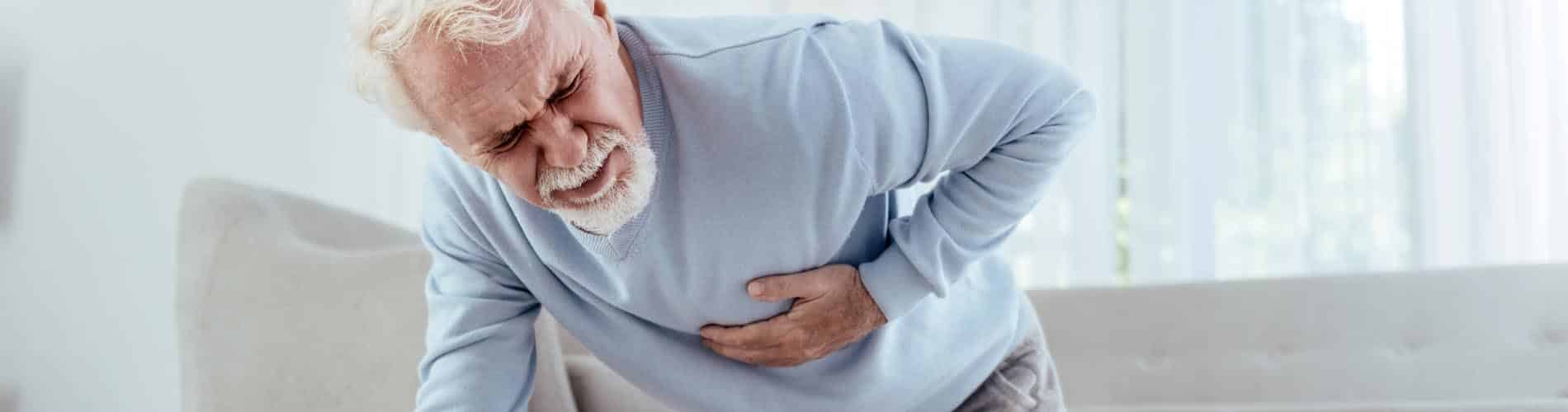 An elderly man with white hair and a beard, wearing a light blue sweater, clutches his abdomen and leans forward in pain, appearing to experience severe discomfort.