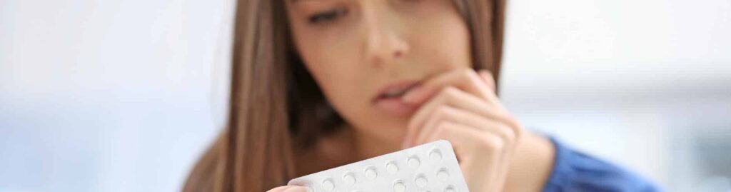 A woman with long brown hair looks thoughtfully at a blister pack of pills, holding it in one hand while resting her chin on her other hand. The background is softly blurred.