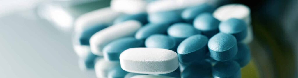 A close-up of a pile of blue and white oval-shaped pills or tablets on a reflective surface, suggesting medical or pharmaceutical use.