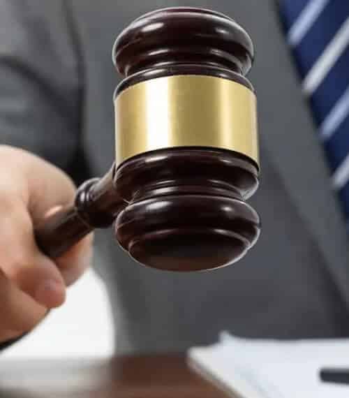 A person in a suit holding a wooden judge’s gavel with a gold band, poised above a desk with documents.