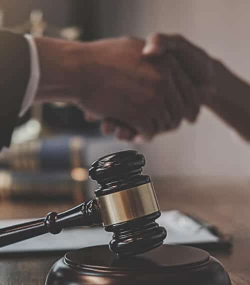 A close-up of a judges gavel resting on a desk, with two people shaking hands in the background, suggesting an agreement or legal settlement.