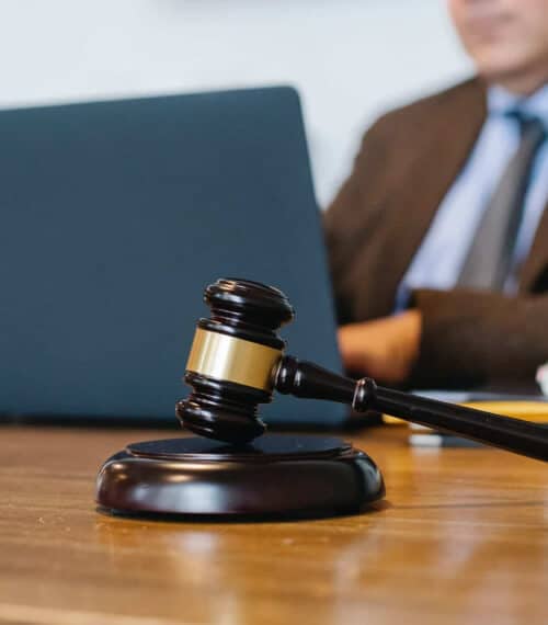 A close-up of a judge’s gavel resting on a wooden desk, with a person in a suit working on a laptop in the blurred background.