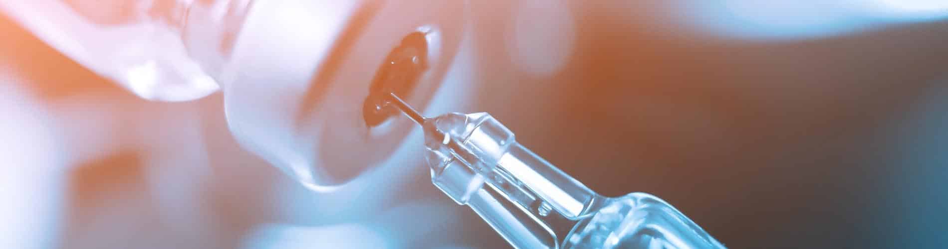 A close-up of a syringe needle inserted into the rubber stopper of a vial, with a blue and orange blurred background.