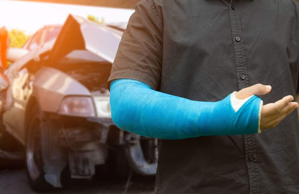A person wearing a blue cast on their arm stands in front of a damaged car, suggesting a recent accident or injury. The person’s face is not visible, and the car appears to have a crumpled front end.