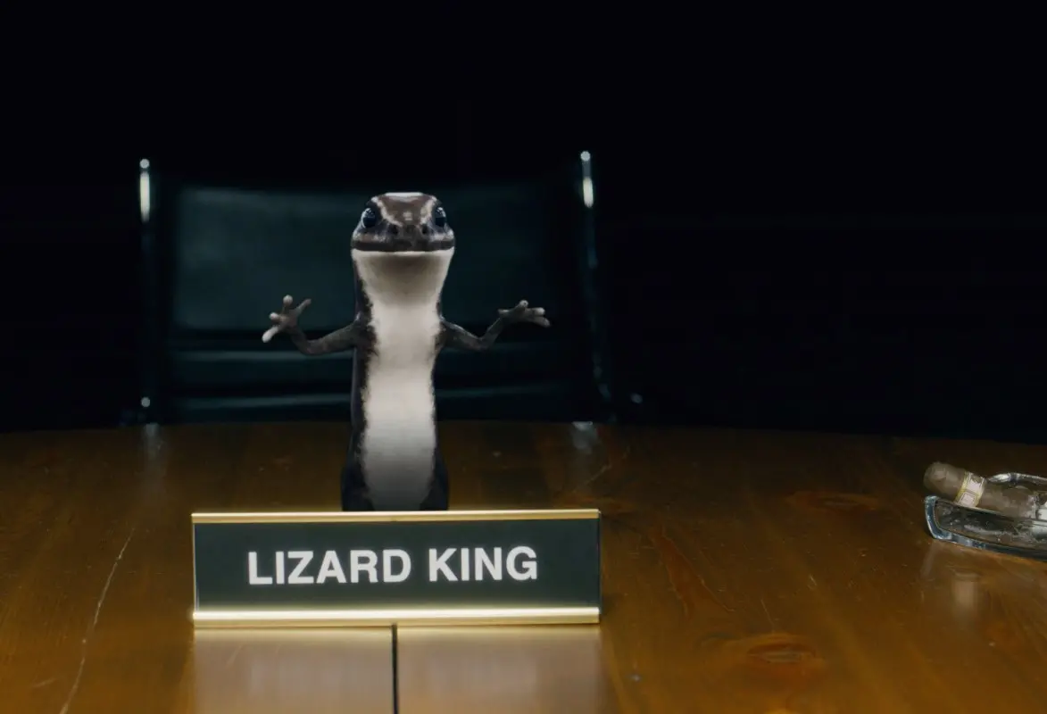 A small lizard stands behind a desk nameplate that reads LIZARD KING, with its front legs raised. A dark chair and a glass bottle lie on the wooden table in a dimly lit room.