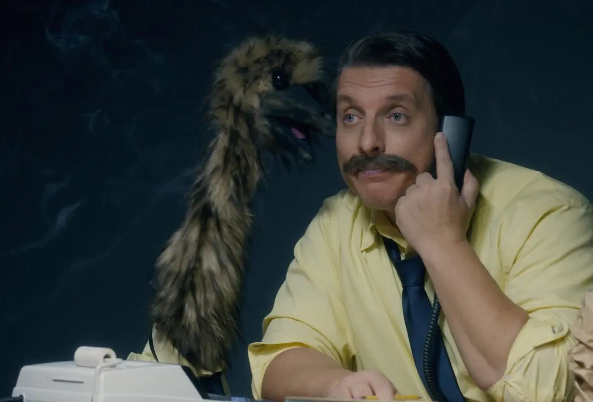 A man in a yellow shirt and blue tie, with a mustache, holds a phone to his ear. Next to him is a furry puppet resembling a bird, looking at him. They are sitting at a desk with a white rotary phone.