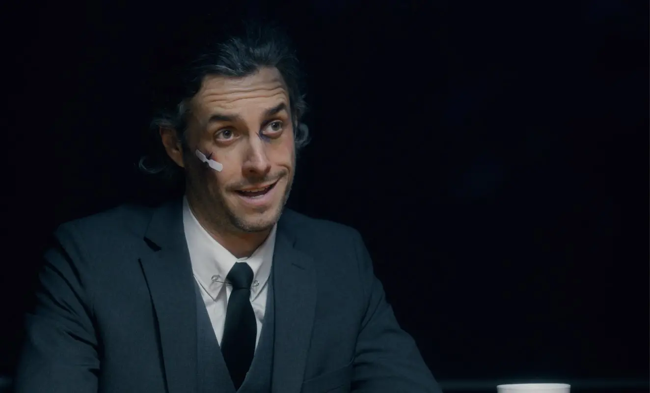 A man in a suit with a small bandage on his left cheek sits at a table, smiling slightly. There is a white cup in front of him, and the background is dark.