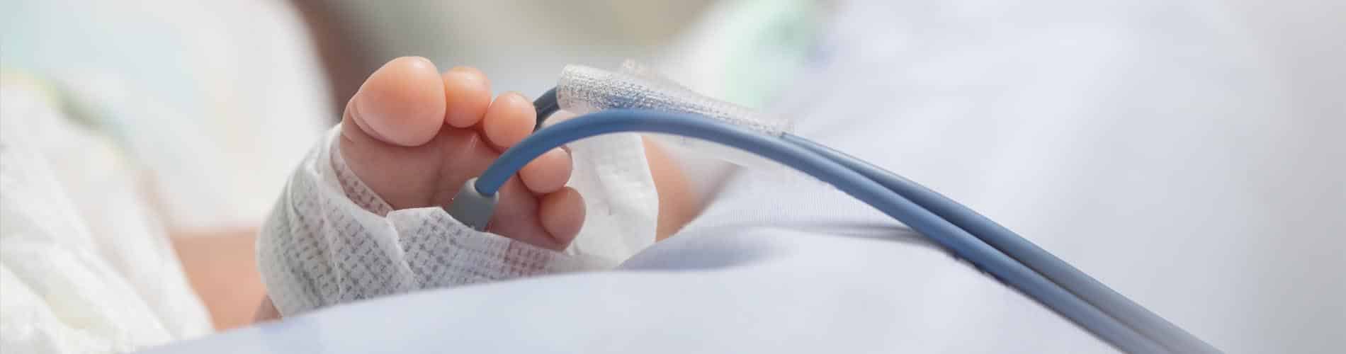 Close-up of a newborn babys foot with medical tape and a blue monitoring tube attached, resting on a white hospital blanket in a clinical setting.