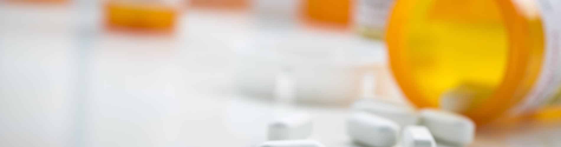 Several white pills are scattered on a smooth surface near an open orange prescription bottle, with more bottles blurred in the background.