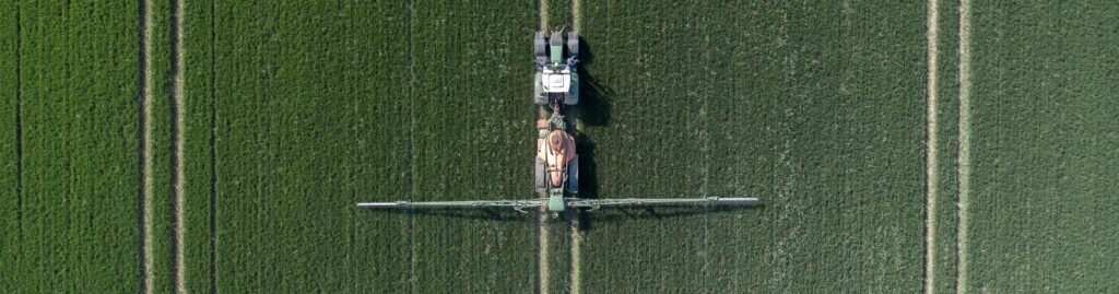 Aerial view of a tractor spraying crops in a green field, creating parallel lines as it moves, with agricultural machinery extended wide across the crop rows.