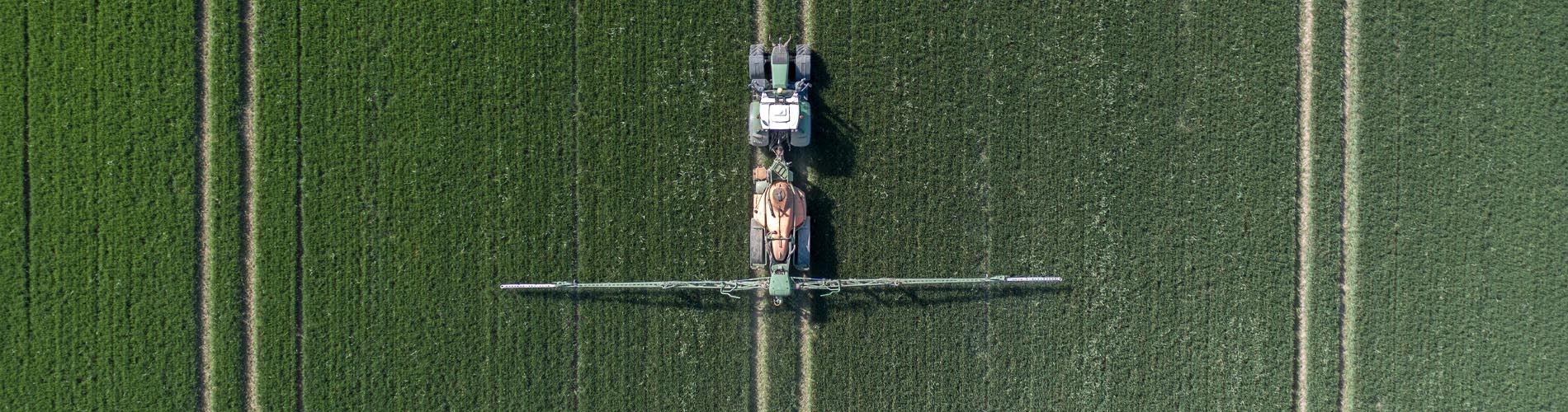 Aerial view of a tractor spraying crops in a green field, creating parallel lines as it moves, with agricultural machinery extended wide across the crop rows.