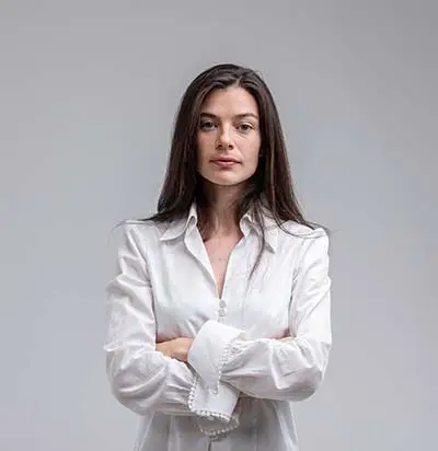 A woman with long dark hair wearing a white button-up shirt stands against a plain light gray background with her arms crossed, looking confidently at the camera.