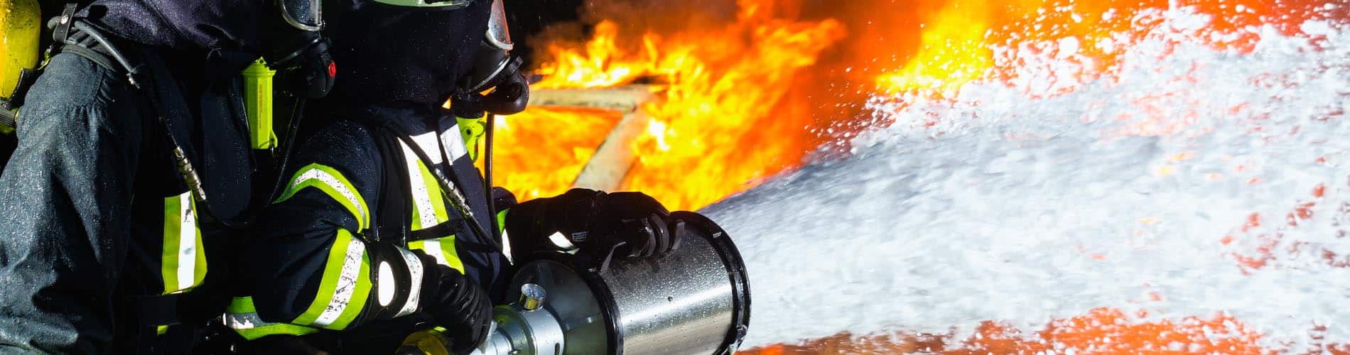 A firefighter in full protective gear sprays foam from a hose to extinguish a large, intense fire with bright orange flames in the background.