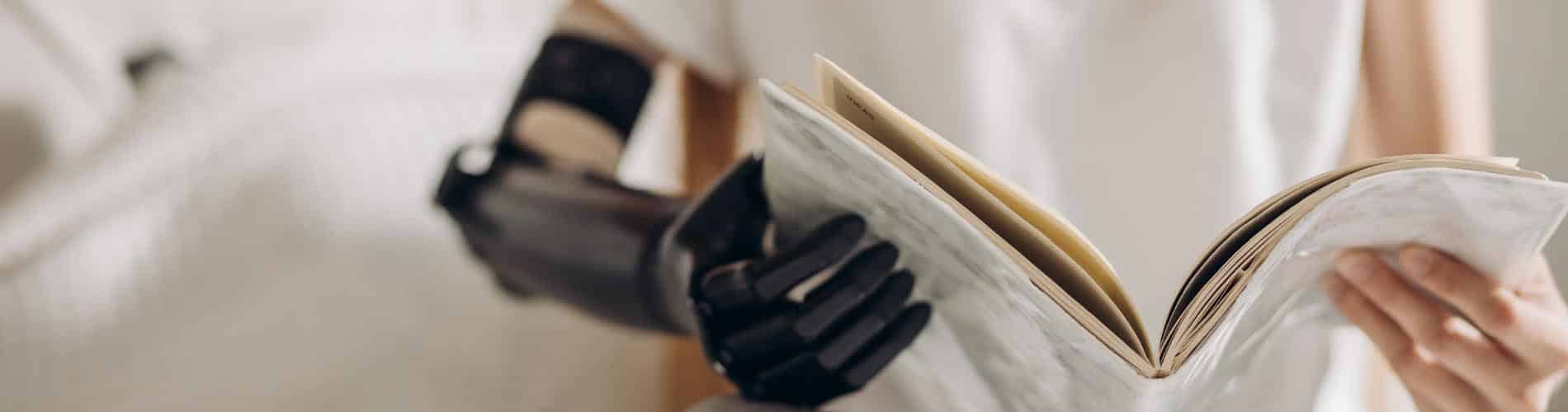 A person with a black prosthetic arm holds and reads an open book, with the focus on their hands and the book’s pages.