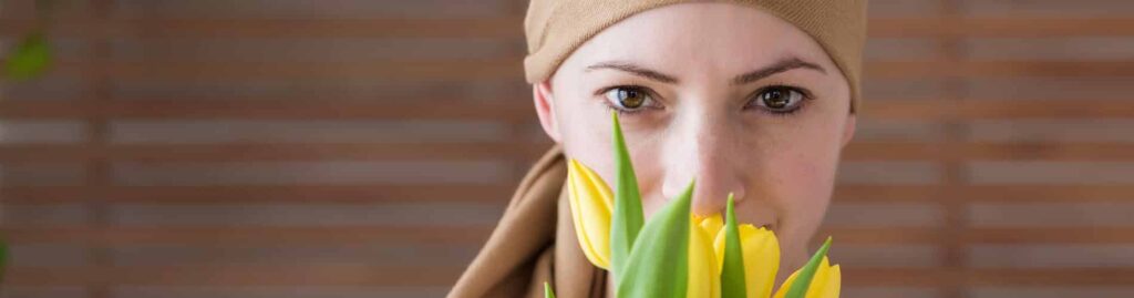 A woman wearing a beige headscarf looks directly at the camera while holding and smelling yellow tulips, with a blurred wooden background behind her.