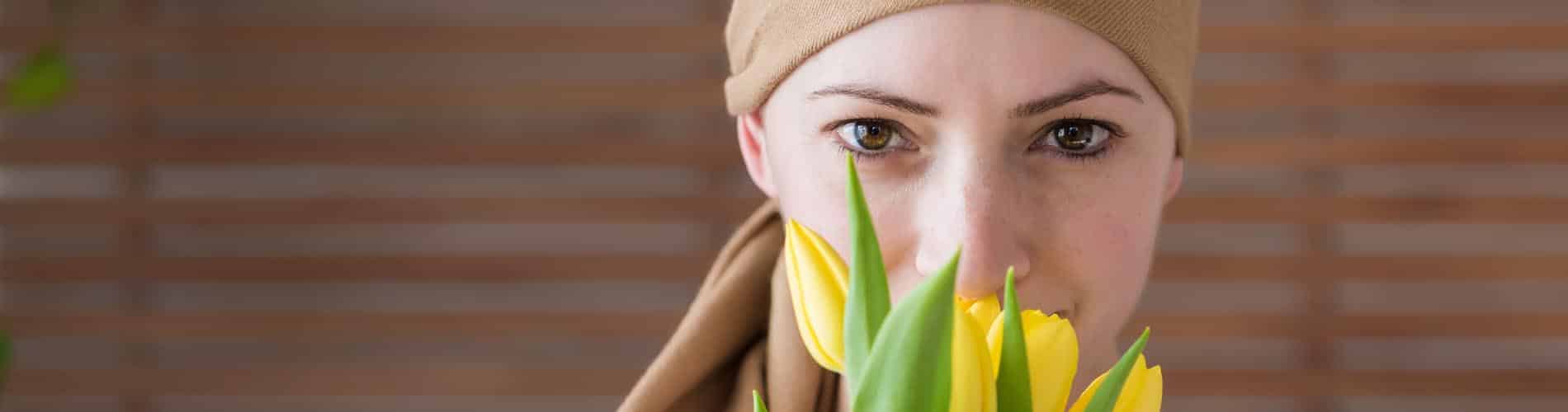 A woman wearing a beige headscarf looks directly at the camera while holding and smelling yellow tulips, with a blurred wooden background behind her.