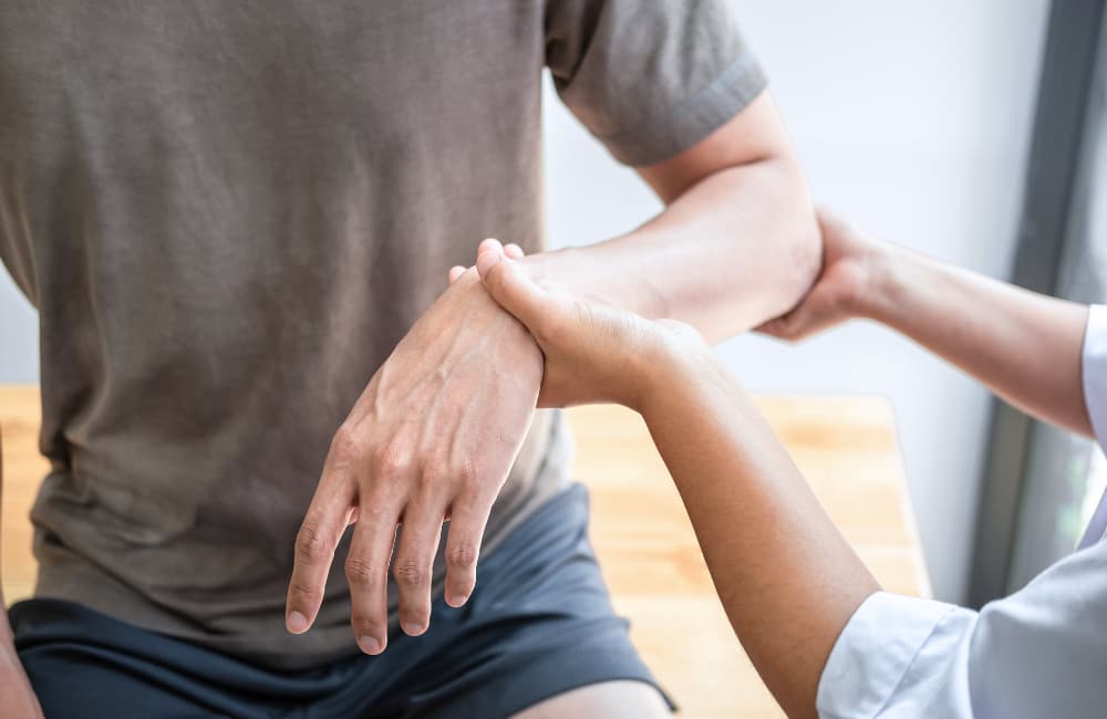 A person wearing a brown shirt and black shorts is having their arm and wrist gently examined or stretched by another person, possibly a healthcare professional, at a wooden table.